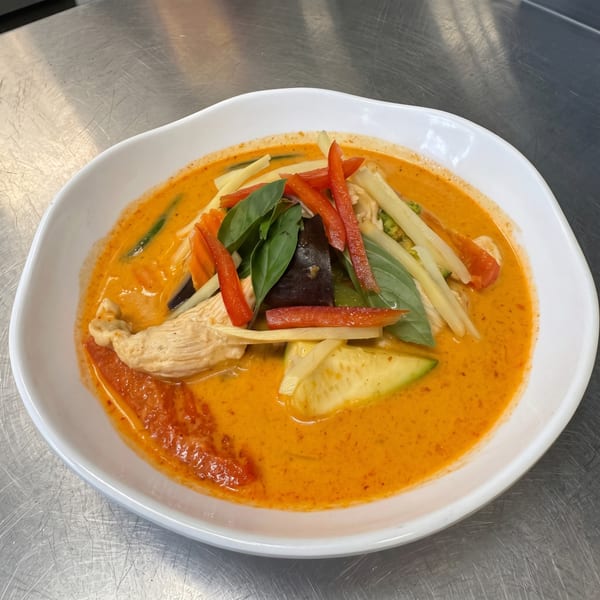 Vibrant red curry with chicken, bell peppers, bamboo shoots, zucchini, eggplant, and basil in a white bowl.