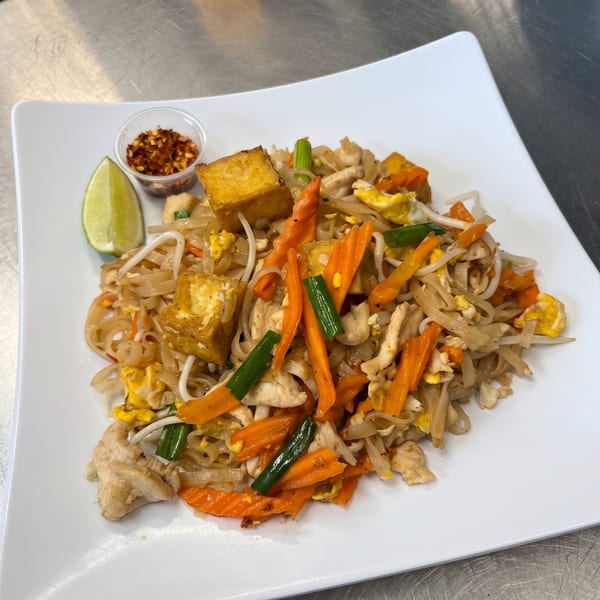 Pad Thai with chicken, tofu, carrots, green onions, egg, lime, and chili flakes on a white plate.